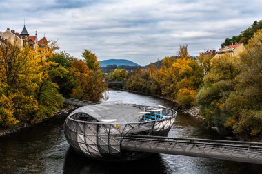 Murinsel Adası 'nın Murinsel Nehri' ndeki güzel manzarası. Arkasında renkli ağaçlar ve dağlar var. Bulutlu bir sonbahar gününde, Graz, Avusturya.