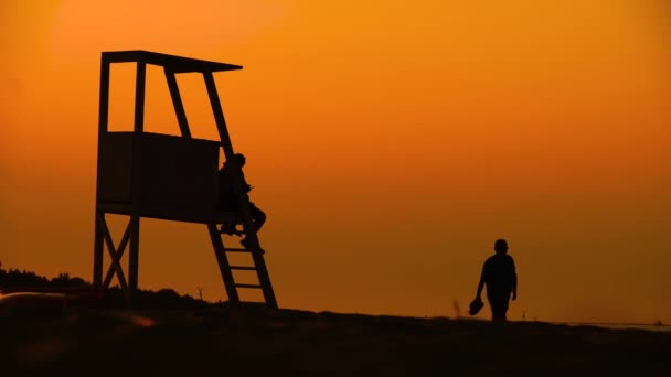 Summertime reisconcept. Donker silhouet, iconische retro houten strandwacht toren tegen zonsondergang oranje hemel. Contrast uitkijktoren omtrek, strand schemering esthetisch. Mensen — Stockvideo