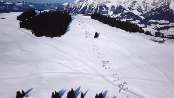 Skigebied bij zonnig weer. Sneeuw op de skipiste van een beboste berg. Veel toeristen in de buurt van de skilift station en cafe. Luchtzicht — Stockvideo