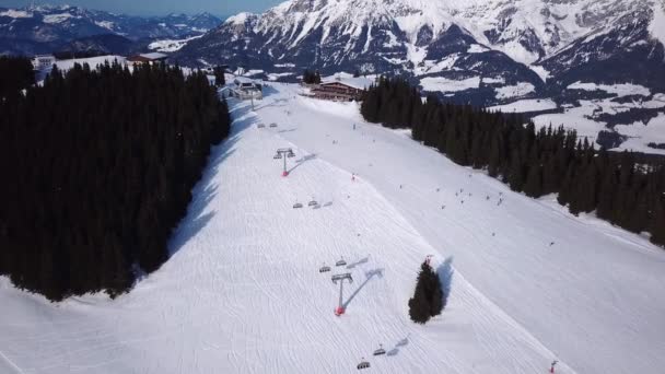 Comprensorio sciistico con tempo soleggiato. Neve sulla pista da sci di una montagna boscosa. Molti turisti vicino alla stazione degli impianti di risalita e caffè. Vista aerea — Video Stock