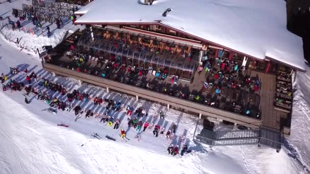 Vídeo aéreo de drones de un restaurante de esquí lleno de gente con terraza. Delantales bar de esquí rodeado de nieve con escenario y música en vivo repleta de esquiadores relajantes. Cabaña de montaña abarrotada de turistas. — Vídeos de Stock