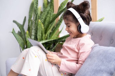 Gülümseyen küçük kız evdeki koltukta kablosuz kulaklıkla tablet kullanıyor..