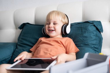 Gülümseyen küçük çocuk evde kablosuz kulaklıkla tablet kullanıyor..