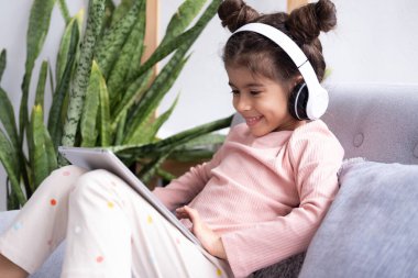 Gülümseyen küçük kız evdeki koltukta kablosuz kulaklıkla tablet kullanıyor.