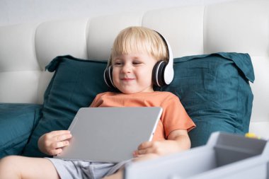 Gülümseyen küçük çocuk evde kablosuz kulaklıkla tablet kullanıyor.