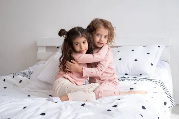 two beautiful diverse kids girls in pajamas hugging on bed in modern bright apartment.