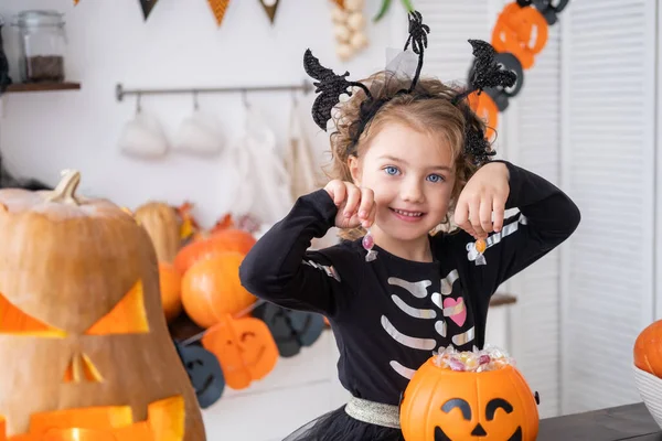 Linda niña en traje de bruja sosteniendo gato de calabaza con caramelos, divirtiéndose en la cocina, celebrando Halloween Imagen De Stock