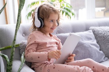 Gülümseyen küçük beyaz kız evdeki koltukta kablosuz kulaklıkla tablet kullanıyor..