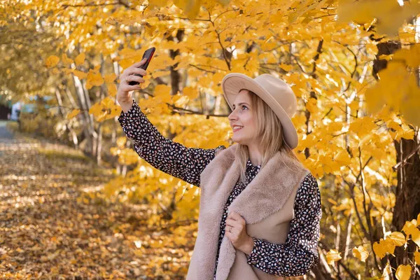 Mulher bonita na roupa na moda e chapéu leva selfie no telefone perto da árvore de outono amarela — Fotografia de Stock