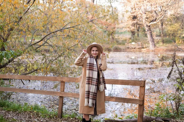 Hermosa mujer joven en abrigo beige, sombrero y bufanda junto al lago en otoño parque en otoño —  Fotos de Stock