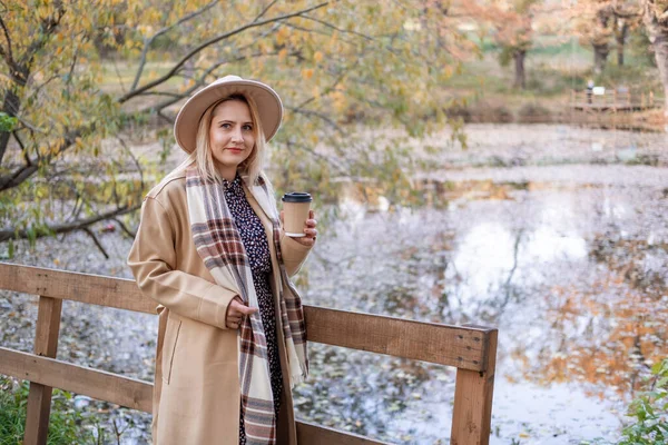 Jovem atraente em casaco bege e chapéu bebe café junto ao lago no parque de outono no outono — Fotografia de Stock