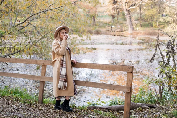 Atractiva joven en abrigo beige y sombrero bebe café junto al lago en otoño parque en otoño —  Fotos de Stock