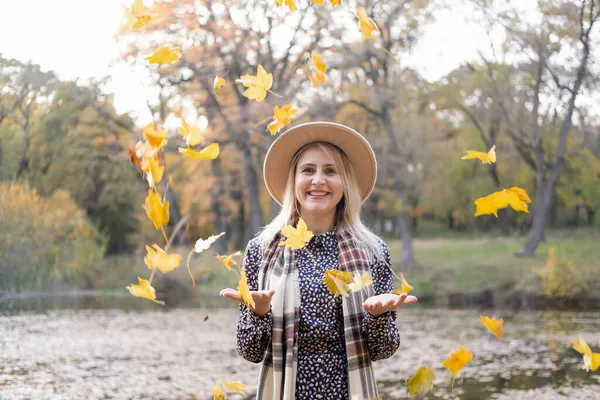 Mulher loira feliz jogando folhas de outono no ar no parque de outono no outono — Fotografia de Stock