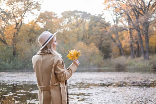 Bela jovem mulher elegante em casaco bege e chapéu detém folhas de outono no parque de outono no outono — Fotografia de Stock