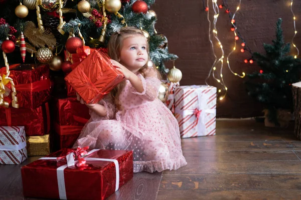 Happy little smiling blond girl in beautiful dress with gift box near christmas tree — Stock Photo, Image