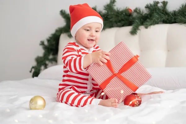 Niña en traje de cuerpo a rayas y Santa sombrero jugando con caja de regalo en el dormitorio decorado de Navidad —  Fotos de Stock