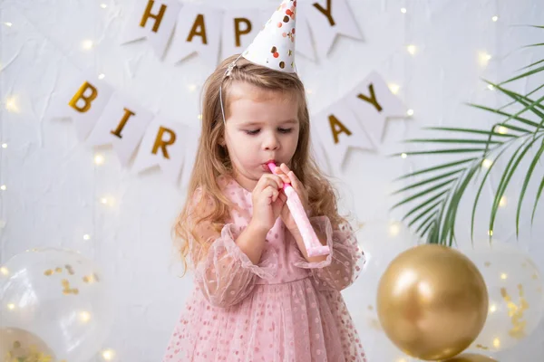 Vtipné dítě dívka v růžových šatech a narozeninové klobouk foukání do píšťalky a slaví své narozeniny — Stock fotografie