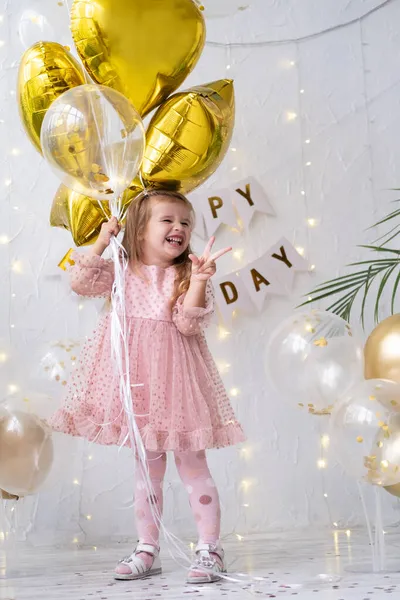 Šťastná malá blondýnka v růžových šatech drží balónky a slaví své narozeniny — Stock fotografie