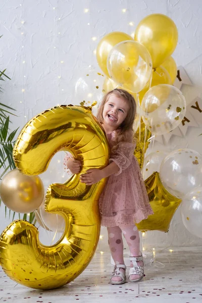 Щаслива маленька блондинка в рожевій сукні тримає велику золоту фольгу номер і цифра 3 — стокове фото