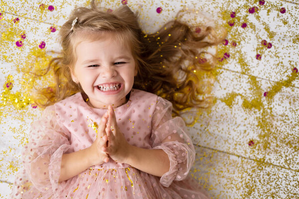 happy child girl in pink dress lying on floor with confetti and celebrating her birthday