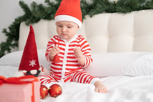 Baby girl in striped bodysuit and Santa hat playing with christmas bell on decorated bed — Stock Photo, Image