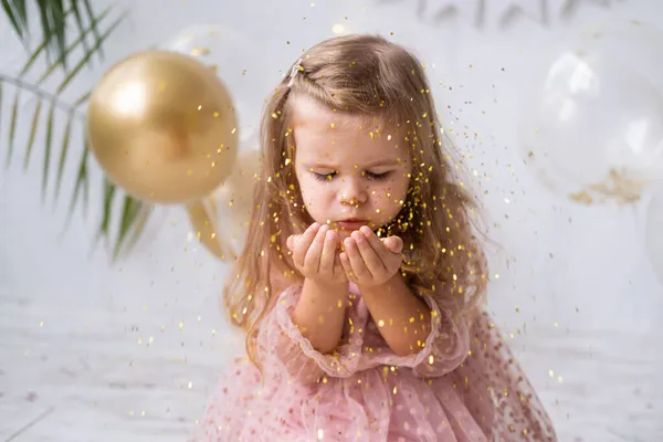 Pembe elbiseli mutlu kız çocuğu konfeti üflüyor ve doğum gününü kutluyor.