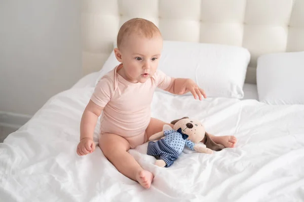 Девочка в светло-розовом костюме, играющая с мягкой игрушкой на белом постельном белье на кровати — стоковое фото