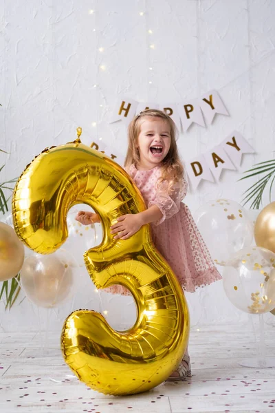 Feliz niña rubia en vestido rosa sosteniendo gran número de globo de lámina de oro y dígito tres 3 Fotos De Stock