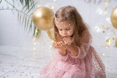 Pembe elbiseli mutlu kız çocuğu konfeti üflüyor ve doğum gününü kutluyor.