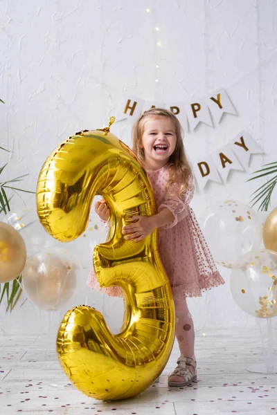 Щаслива маленька блондинка в рожевій сукні тримає велику золоту фольгу номер і цифра 3 — стокове фото