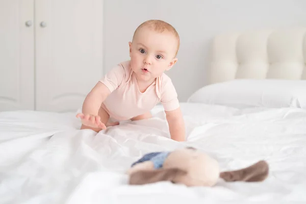 Baby girl in light pink bodysuit playing with soft toy on white bedding on bed — Stock Photo, Image