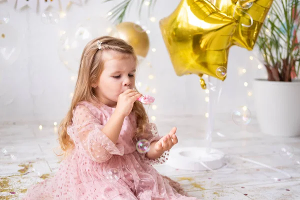 Щаслива дитина дівчина з довгим волоссям в рожевій сукні дме в свист і святкує її день народження — стокове фото