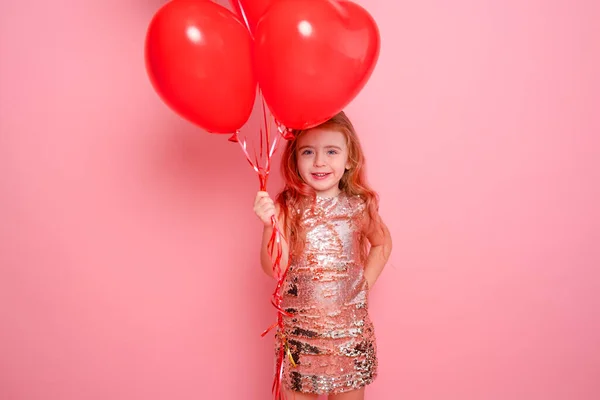 Mooi kind meisje in jurk met pailletten met romantische rode hart ballonnen op roze achtergrond — Stockfoto