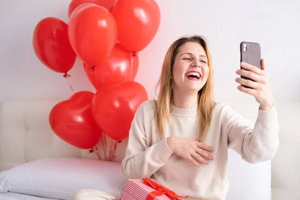 Красивые женщины в повседневной уютной одежде с помощью телефона на кровати празднуя День Святого Валентина — стоковое фото