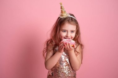 Pembe arka planda doğum günü şapkalı komik kız donut yiyor.