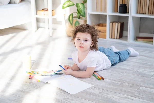 Кудрявая девочка рисует цветными маркерами, лежащими на полу в гостиной дома — стоковое фото