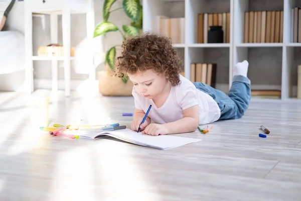 Кудрявая девочка рисует цветными маркерами, лежащими на полу в гостиной дома — стоковое фото