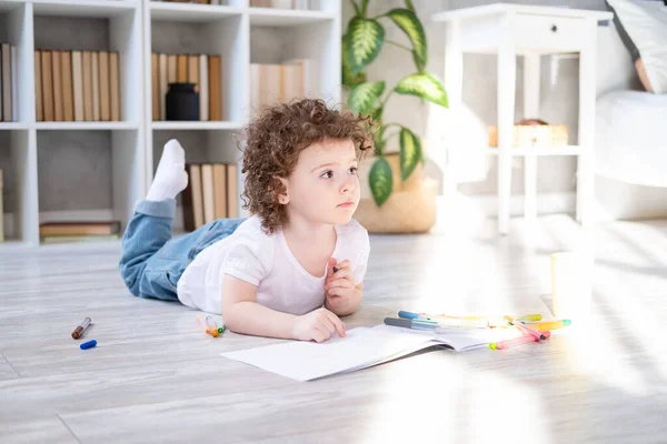 Criança encaracolado menina desenho com marcadores coloridos deitado no chão na sala de estar em casa — Fotografia de Stock