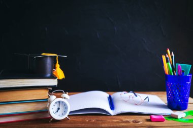 Masaüstünde kitaplar, yazma malzemeleri, gözlükler, defter ve çalar saat var. Siyah arka plan. Öğrenme ve kendini eğitme konsepti. Seçici odak.