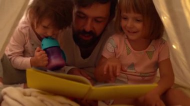 Baba ve küçük kızları çadırda kitap okuyorlar. 