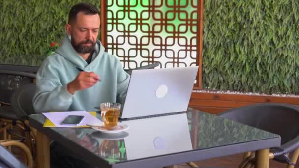 Человек с ноутбуком в кафе пишет заметки — стоковое видео