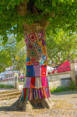 Parkta örülmüş renkli kapaklı bir ağaç gövdesi. Sıcaklık, koruma ve dekorasyon için yamalı örgü örülmüş ağaç..
