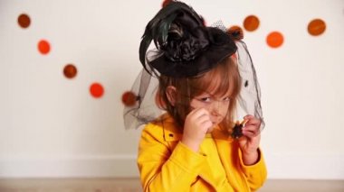 Cadı şapkalı gülümseyen kız jöle örümcek şeklinde şeker yiyor ve oynuyor.