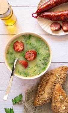 Portuguese style soup called Caldo Verde, bread, Cabbage julienne, oil, and chorizo sausage on a white wooden Table. Flat lay