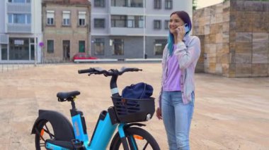 Happy hipster female talking on the phone, standing near an electric bicycle on the urban street. ECO concept. Eco-friendly transport. 