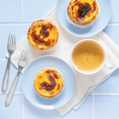 Traditional Portuguese egg tarts dessert Pasteis Pastel de nata or Pastels de Belem with a cup of coffee and forks on the blue tile. Flat lay