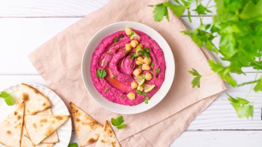 Beetroot hummus decorated with chickpeas on pink towel, pita bread, parsley on the white wooden table background