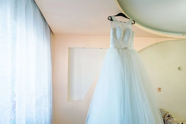 The perfect wedding dress with a full skirt on a hanger in the room of the bride with blue curtains