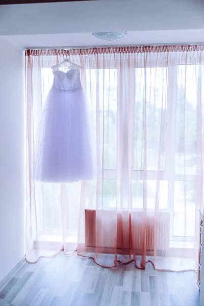 The perfect wedding dress with a full skirt on a hanger in the room of the bride with blue curtains