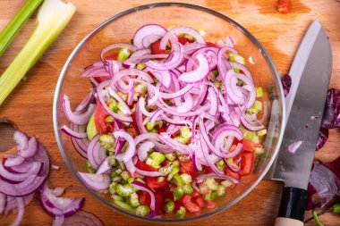 Cooking vegan salad - chopped red onions, cucumbers, tomatoes and celery in a glass bowl, on a table with a knife and ingredients, top view